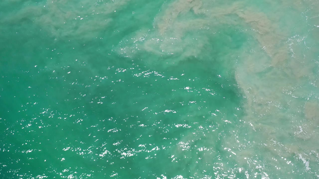 Overhead Los Cabos water frame with turquoise ripples, scattered white glints, a cloudy sand patch beneath the surface, and no visible people.