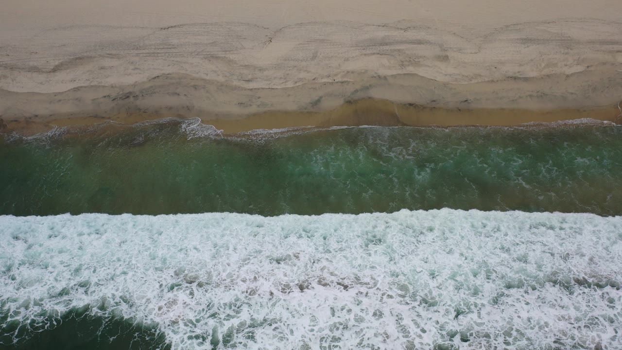 Overhead view of white surf reaching a tan Los Cabos beach with darker wet sand bands and no visible people.