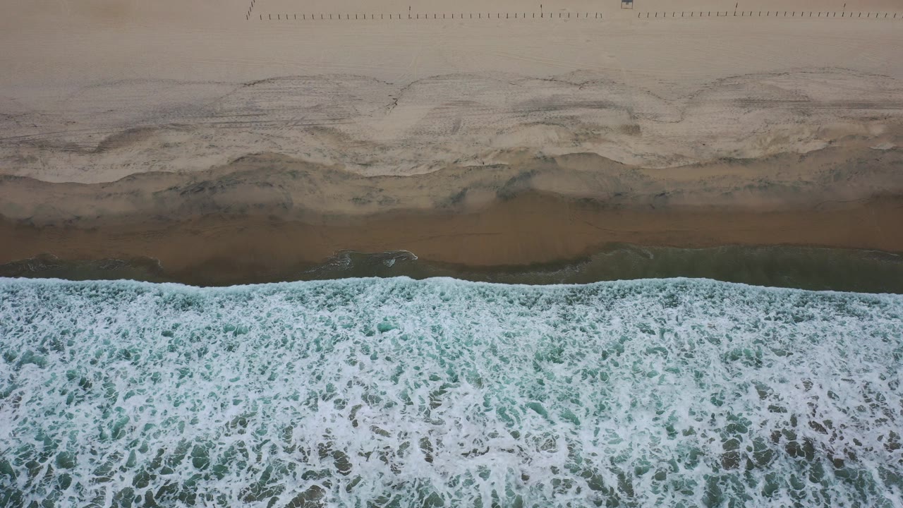 Overhead view of white surf rolling toward brown sand in Los Cabos, Mexico, with darker wet sand above the waterline and no visible people.