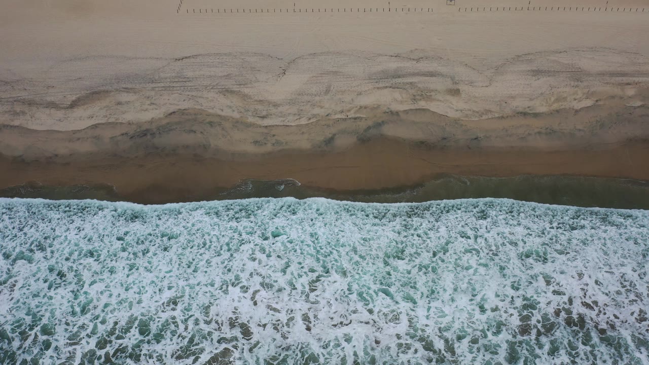 A broad band of white ocean foam spreads across green water beside the smooth brown shoreline in Los Cabos, Mexico, with no visible people.
