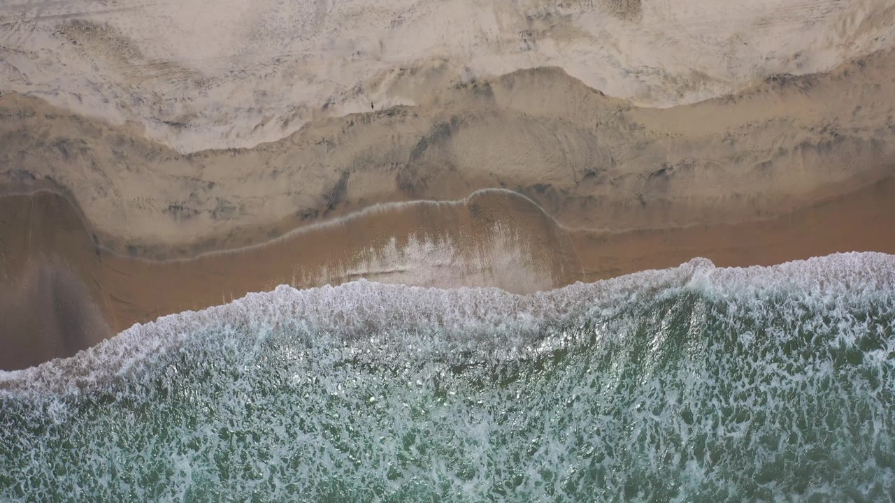 Aerial Los Cabos shoreline frame with white wave foam along the lower edge, layered brown wet sand, pale dry sand, and no visible people.