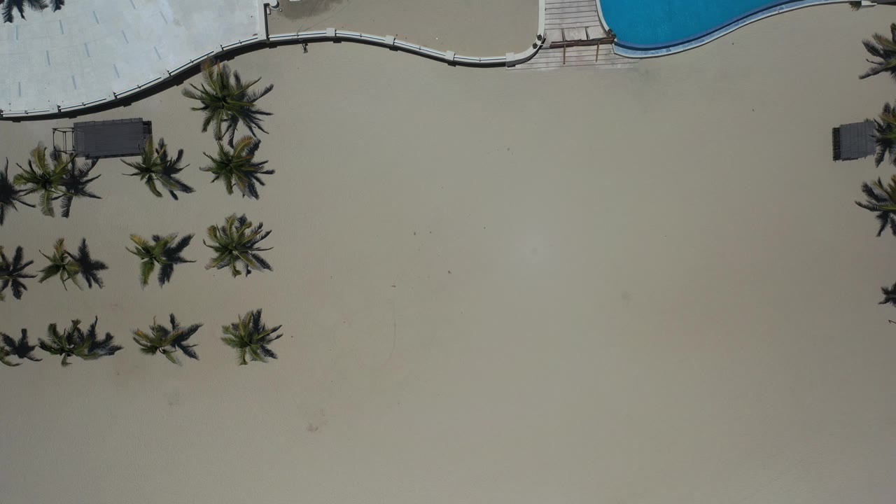 Overhead resort view with palms, sand, decking, and part of a blue pool.