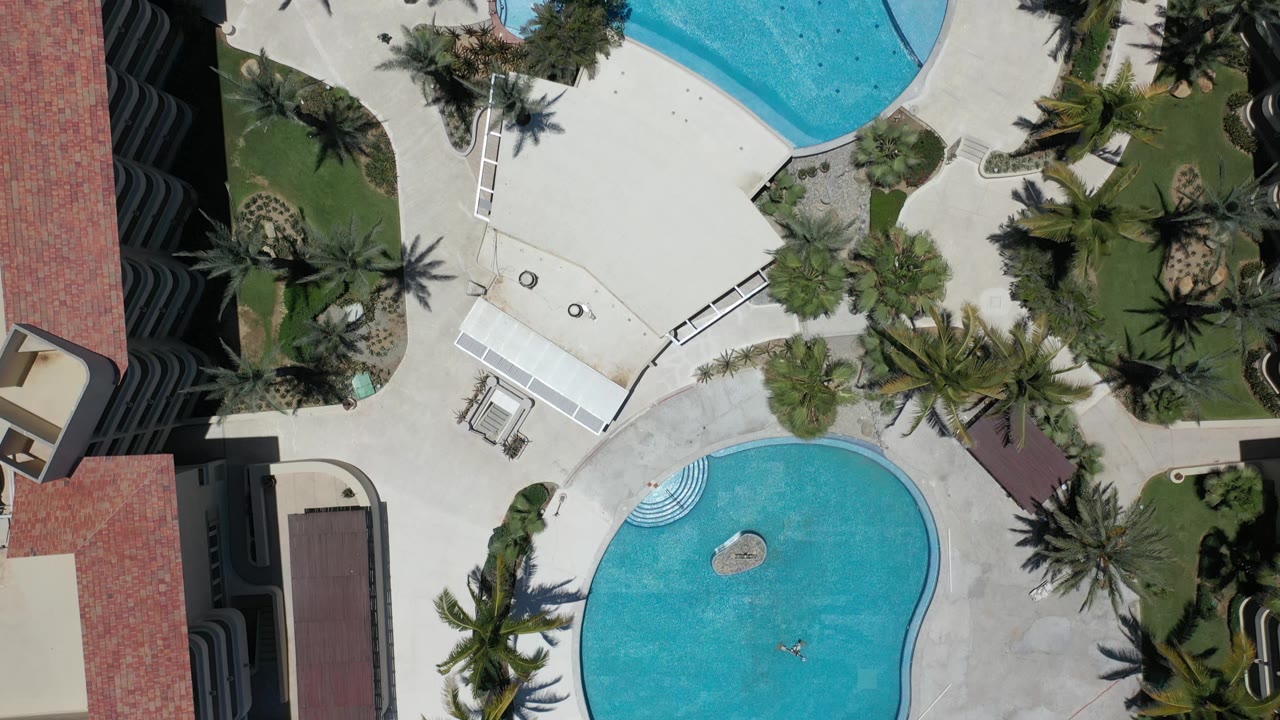 Top-down Los Cabos resort view with curved turquoise pools, planted lawn islands, palm shadows, red roof sections, dark balcony lines, and no visible people.