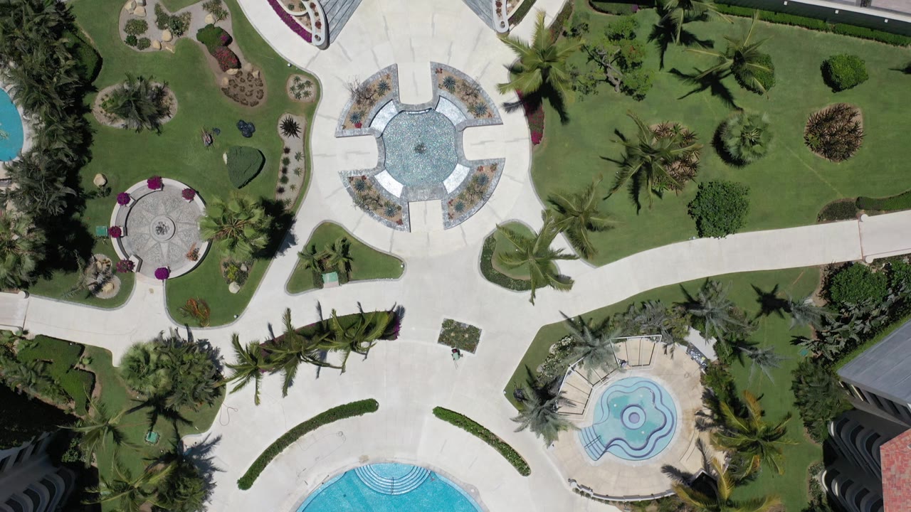 Top-down Los Cabos frame with manicured grass, palm shadows, a round plaza-like feature, pale walkways, partial pool shapes, and no visible people.