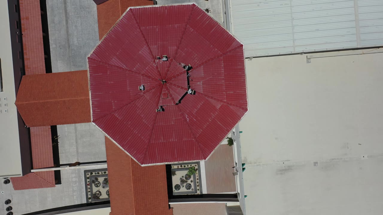 A near-vertical aerial frame in Los Cabos, Mexico centers the red roof shape with light roof panels and paved service areas around it, with no visible people.