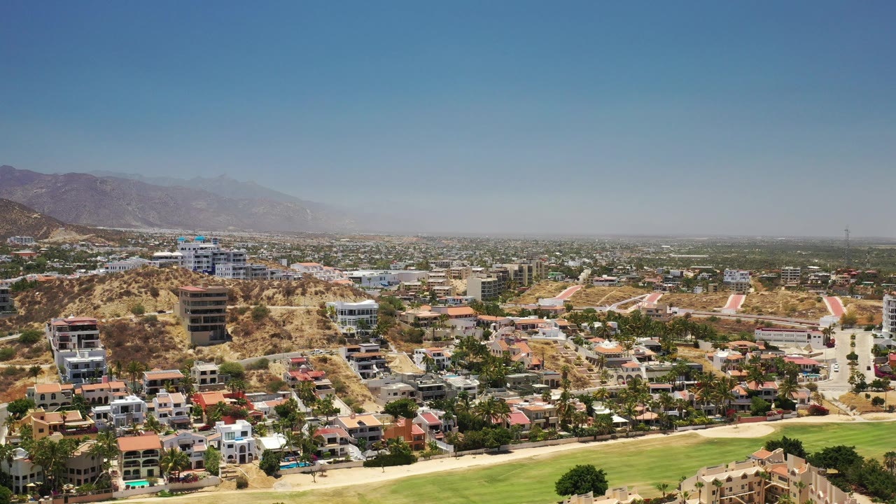 Wide aerial view over Los Cabos with hillside homes, a green landscaped foreground, distant urban spread, hazy mountains, and no visible people.