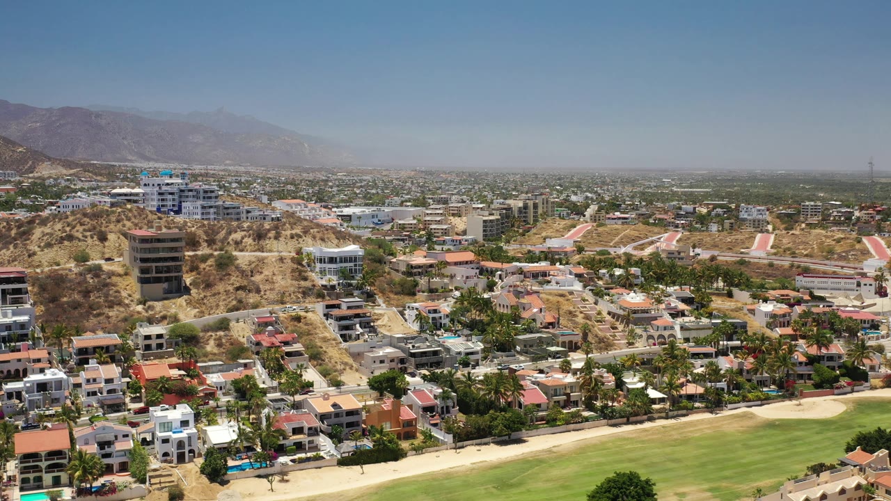 Los Cabos aerial view with a wide green foreground, clustered homes, hillside roads, low-rise construction, distant mountains, and no visible people.