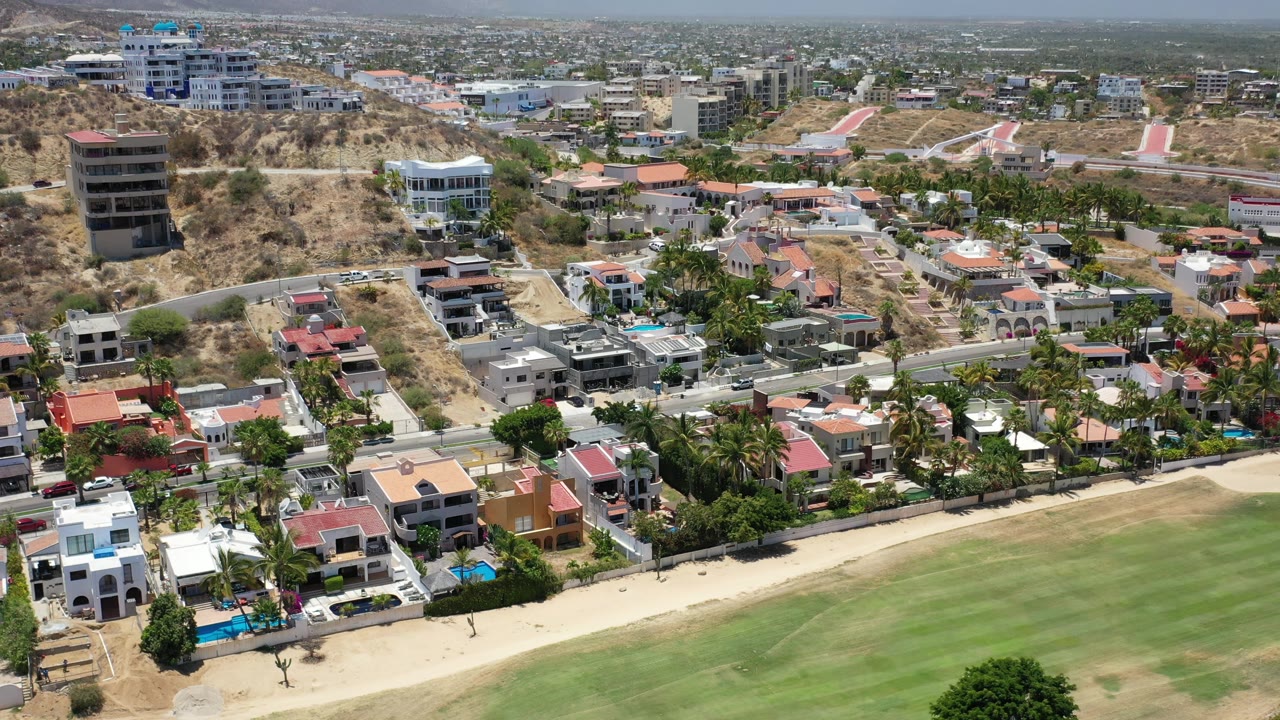 Los Cabos aerial frame looking over a foreground lawn toward a row of homes with pools, palm trees, a paved road, dry slopes, and distant buildings under hazy daylight.