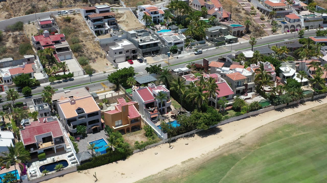High-angle aerial view of houses, road, palms, pools, and green open space.
