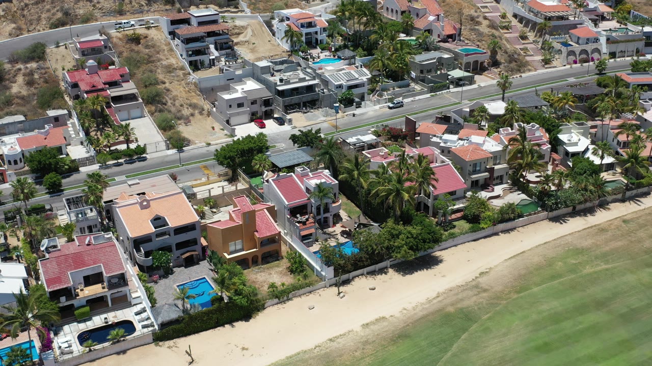Aerial frame in Los Cabos, Mexico, with the road crossing near the top, clustered homes and palms below, a blue pool at lower left, green open space at lower right, and no visible people.