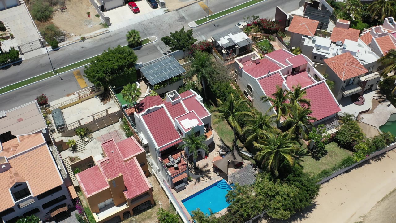 Overhead Los Cabos frame with a central red-roofed house, palms crossing the lower half, a turquoise pool at the lower edge, and no visible people.