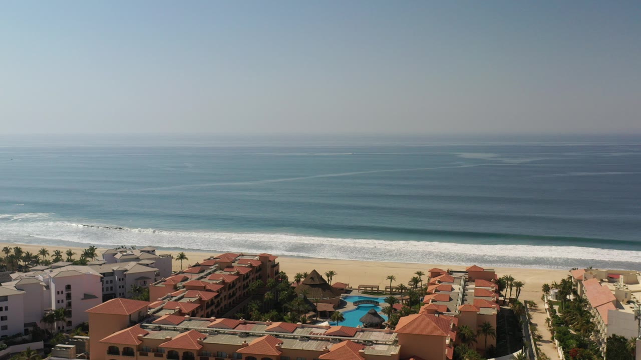 Opening aerial frame over Los Cabos with red tile resort roofs and blue pools below a sandy beach, white surf, and a hazy ocean horizon, with no visible people.