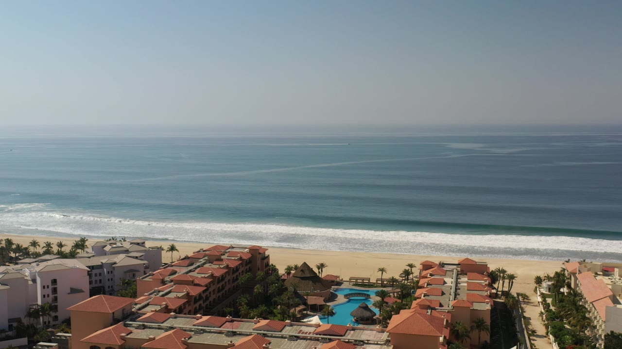 Beachfront hotels and pools below a wide ocean view with waves breaking on sand.