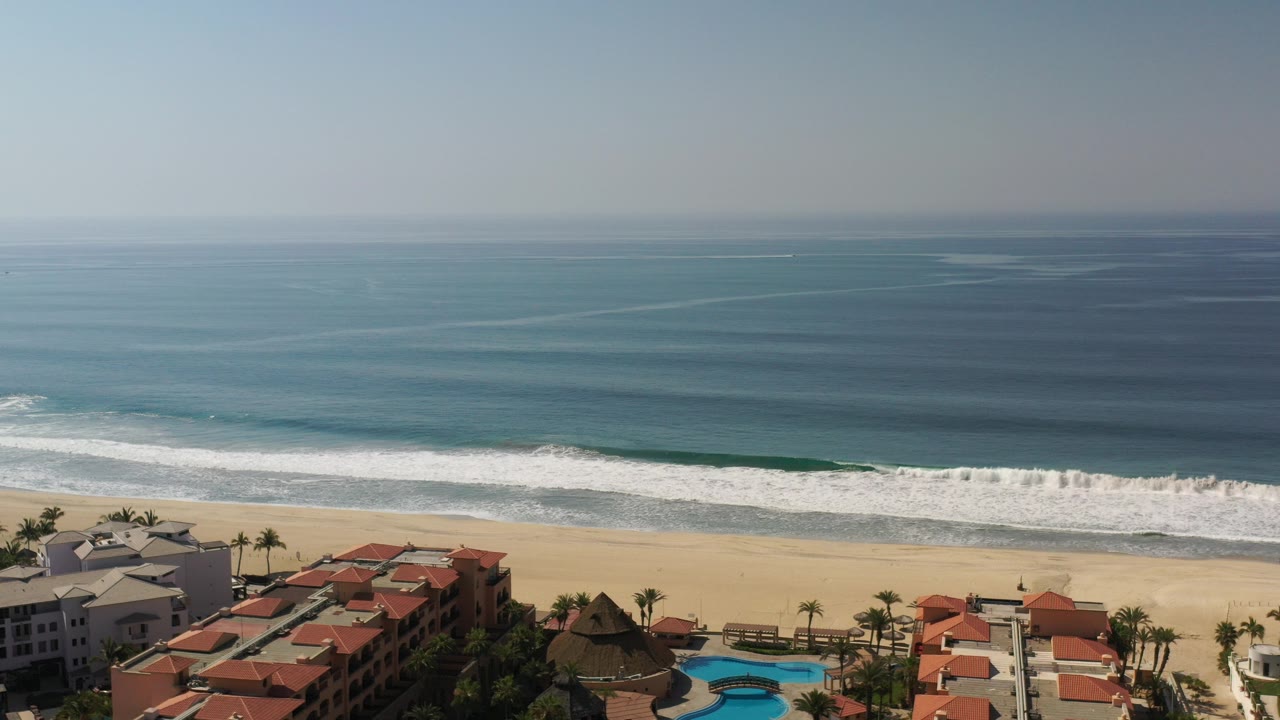 Aerial view of blue ocean, white surf, sandy beach, and red-roofed beachfront buildings.