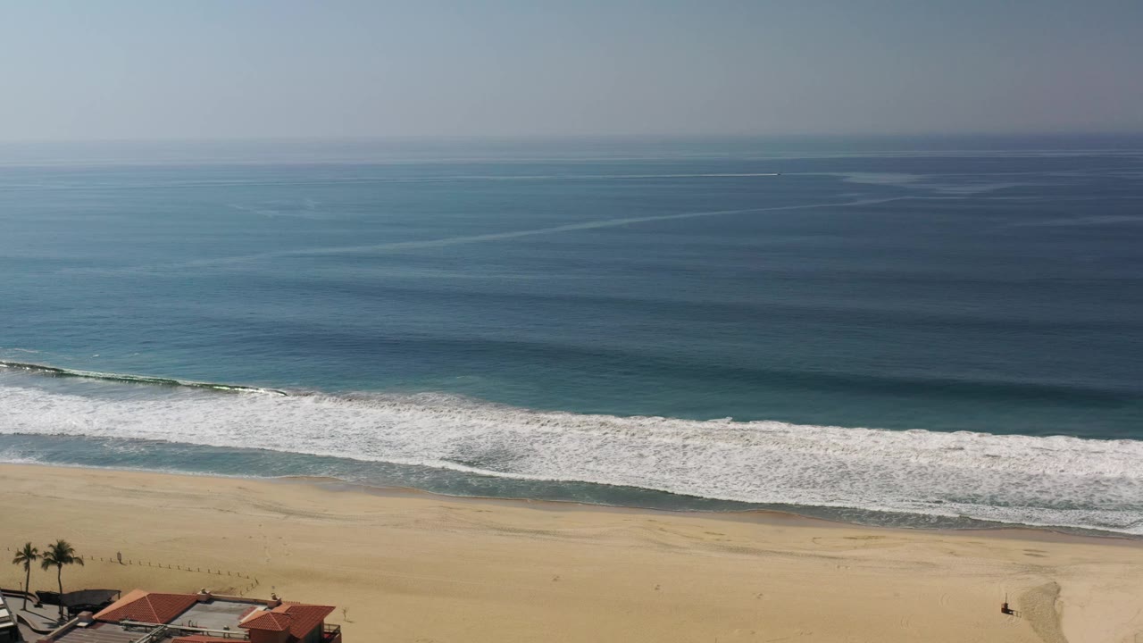 High view of the Los Cabos shoreline with broad blue water, a white foam line, open sand, a few roof edges at the bottom, and no visible people.