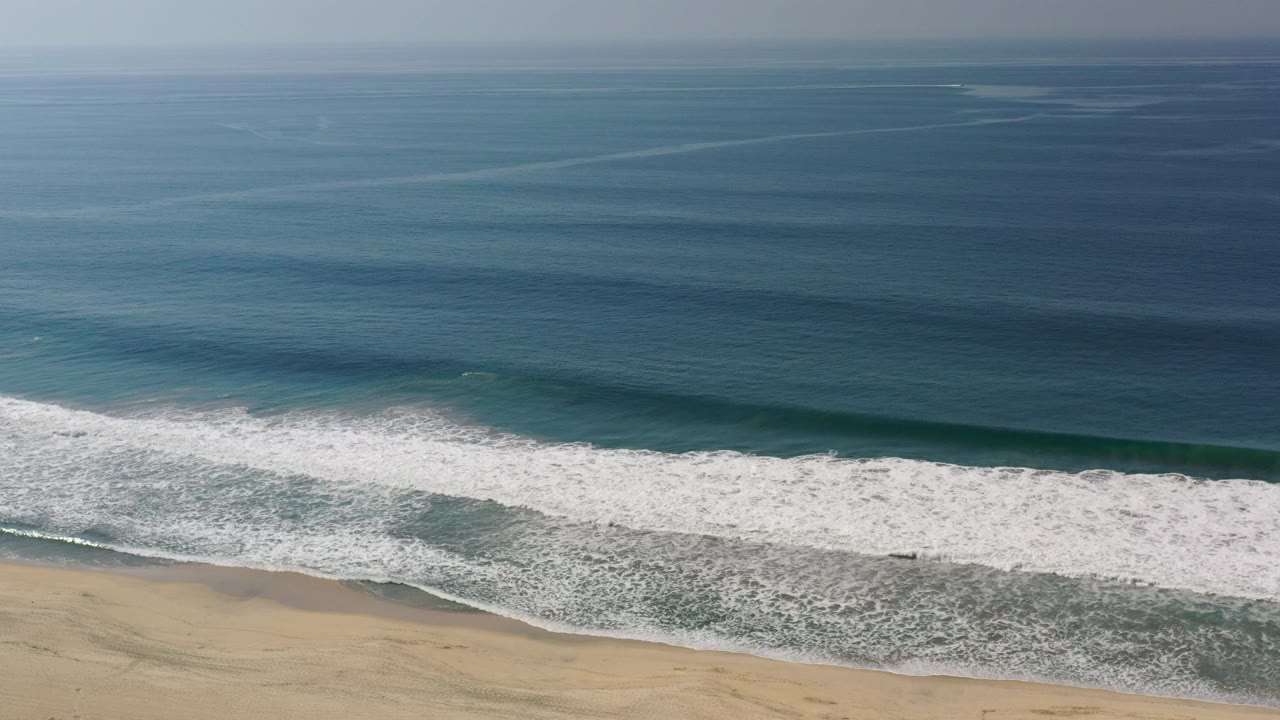 Aerial view in Los Cabos, Mexico, with deep blue water above, a wide white surf band crossing the frame, pale sand at the bottom, and no visible people.