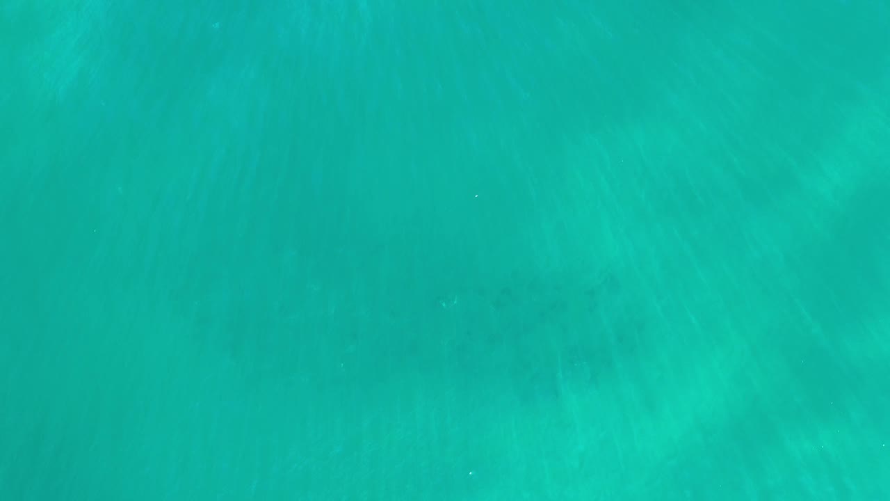 Aerial frame of blue-green Los Cabos water with soft darker patches below the surface and no visible people.