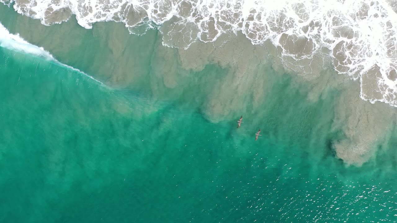Aerial Los Cabos surf frame with teal-green water, pale sand clouds beneath the surface, and white foam curling across the upper shoreline, with no visible people.
