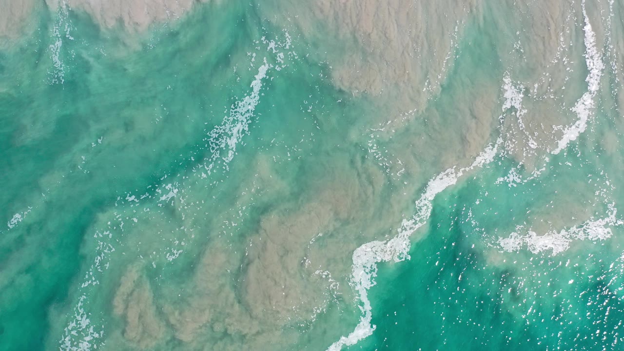 Overhead view of teal ocean in Los Cabos, Mexico, with white foam streaks crossing gray-brown shallow water and no visible people.