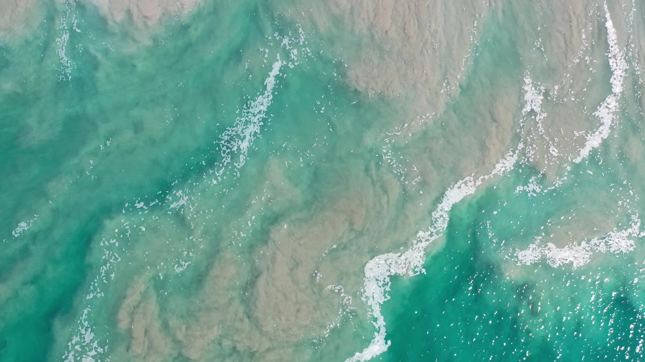 Top-down ocean surface with teal water, foam, and pale gray areas.