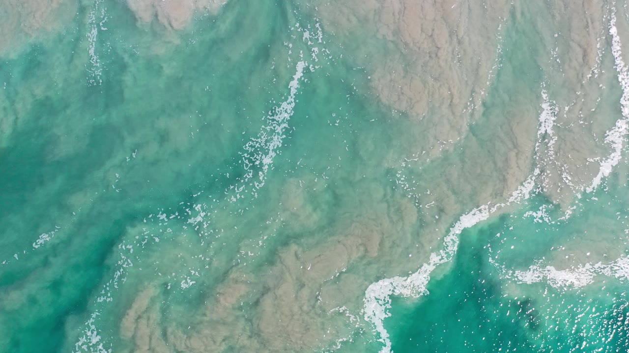 Top-down view of green ocean water in Los Cabos, Mexico, with a curved white foam line over a pale sandy seabed and no visible people.
