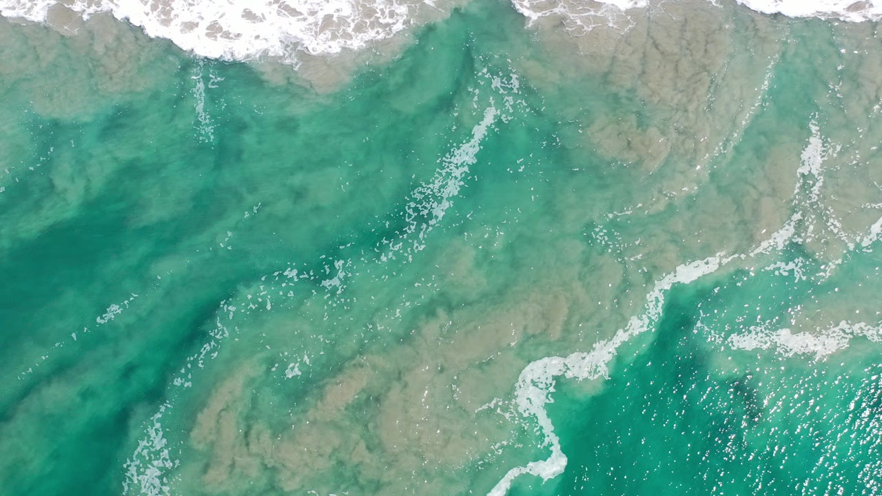 Overhead view of green-blue ocean water in Los Cabos, Mexico, with white surf foam along the top edge and no visible people.