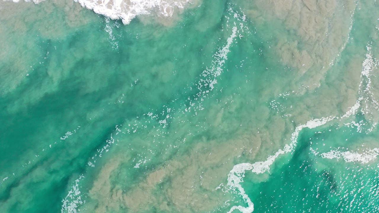 Overhead view of turquoise ocean water in Los Cabos, Mexico, with white surf along the top edge, tan submerged rock shapes, bright surface sparkle, and no visible people.