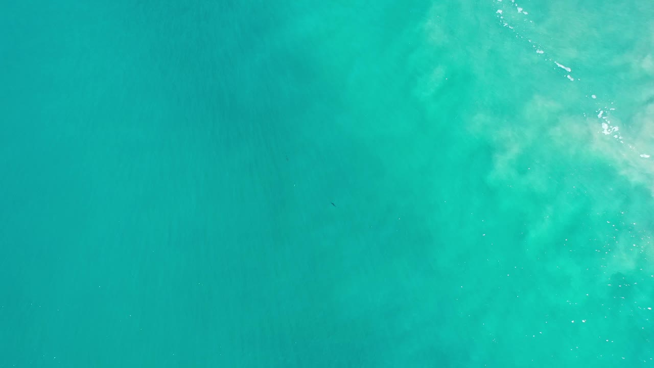 Turquoise water seen from above with pale foam and sand texture at the right edge.