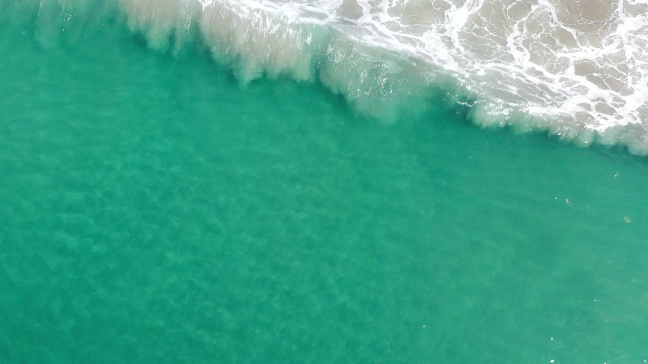 Overhead view of clear green water in Los Cabos, Mexico, with white surf beginning to spread along the top edge and no visible people.