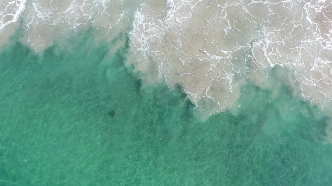 Overhead view of shallow turquoise sea in Los Cabos, Mexico, with white surf spreading across pale sand at the top and no visible people.