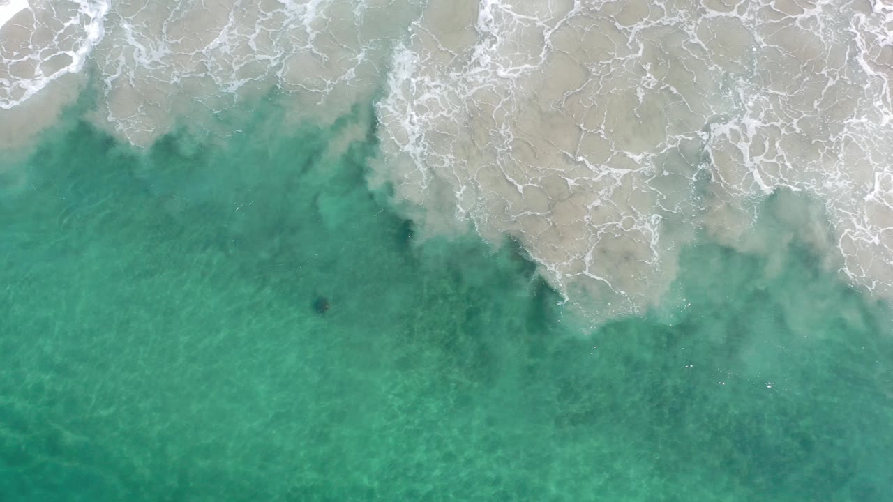 Aerial surf foam spreading across pale shallows.
