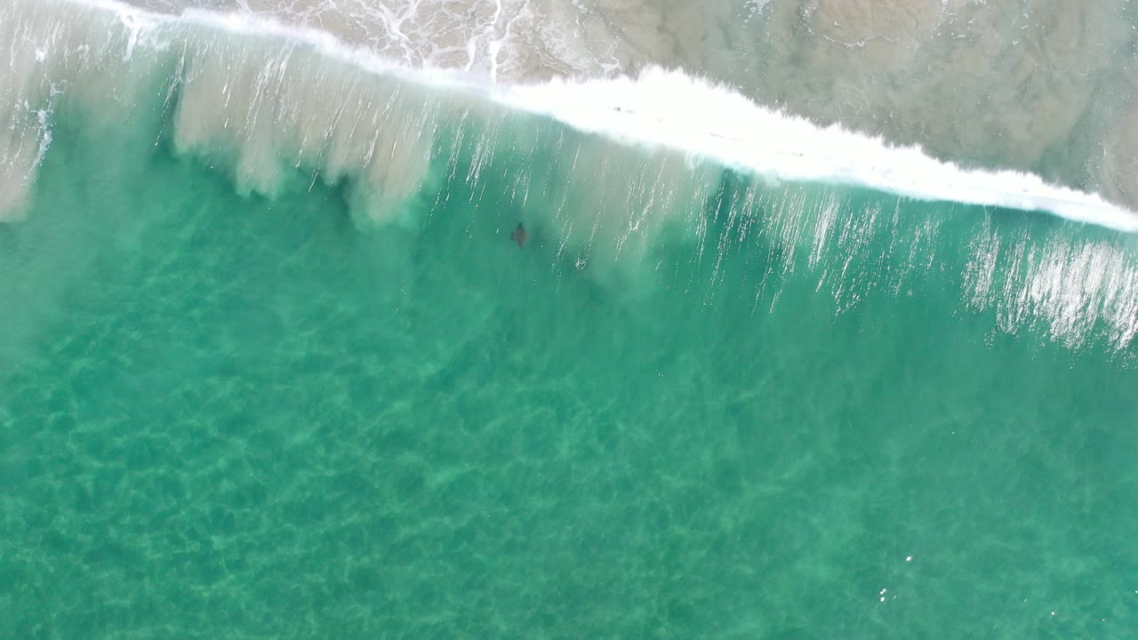 Overhead view in Los Cabos, Mexico, showing turquoise water, pale sand beneath the surface, and white surf entering from the top edge with no visible people.