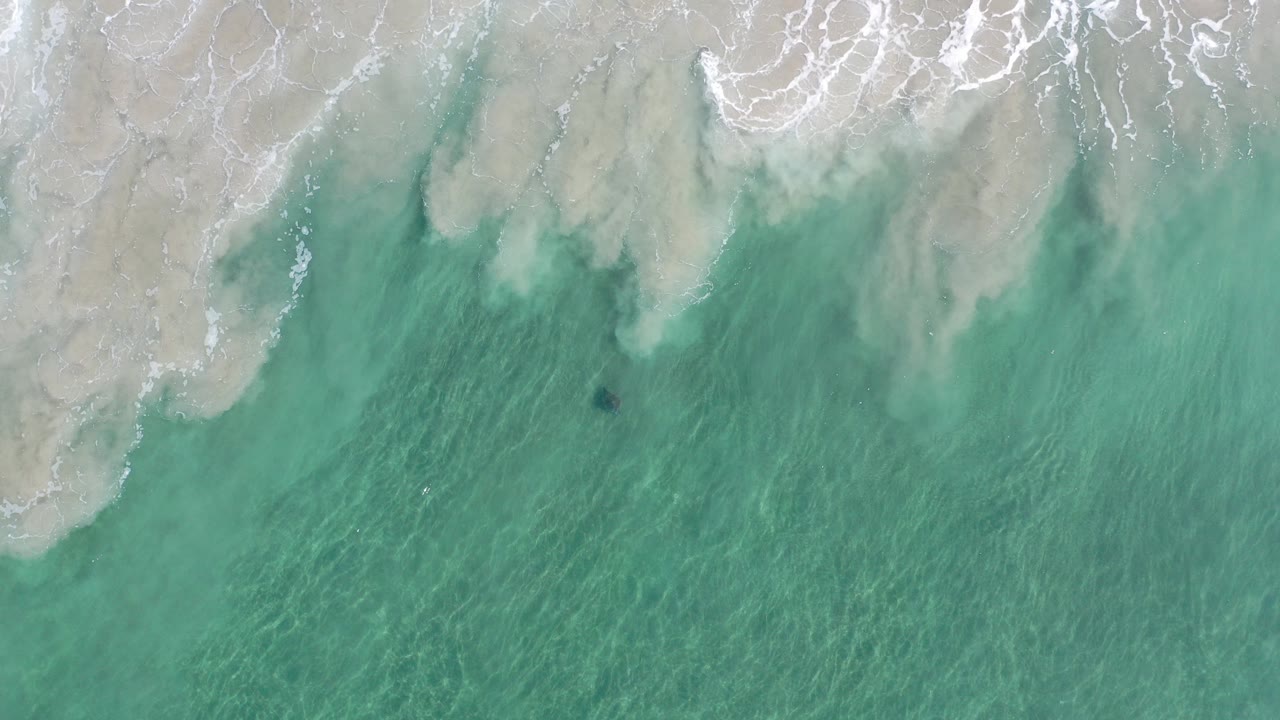Top-down view in Los Cabos, Mexico, with turquoise water below pale wet sand and white foam near the upper edge, no visible people.