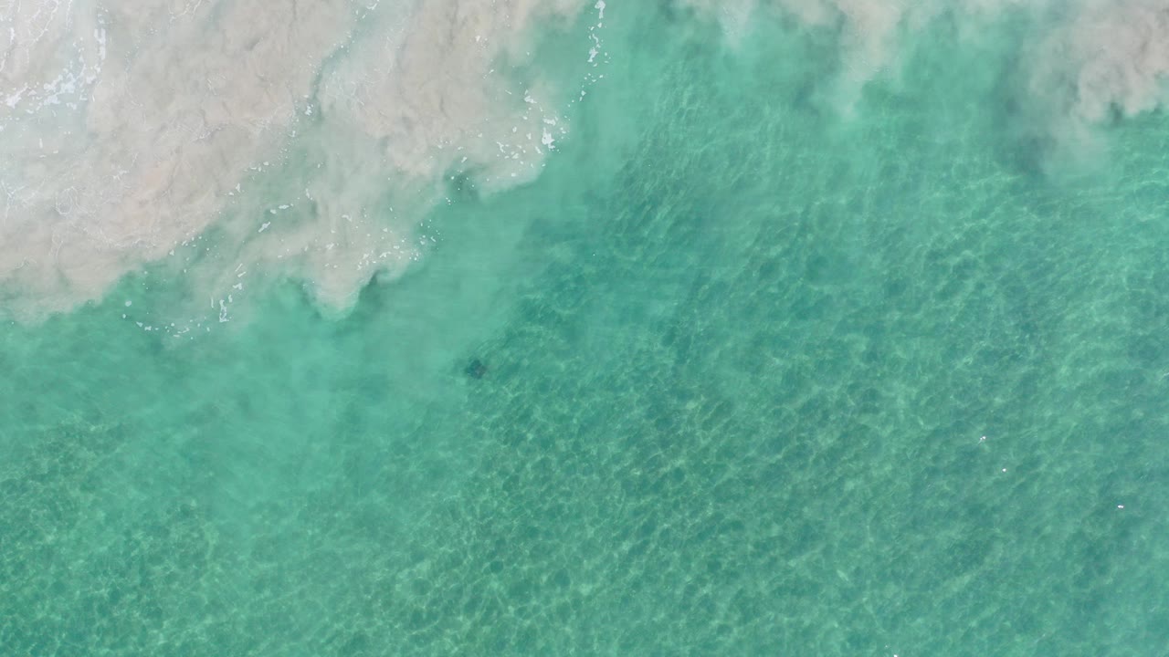 Overhead view of clear turquoise water meeting pale sand in Los Cabos, Mexico, with soft foam near the top edge and no visible people.