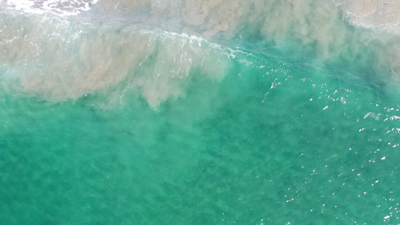Overhead view of translucent turquoise water in Los Cabos, Mexico, with a pale foamy wave band at the top and no visible people.