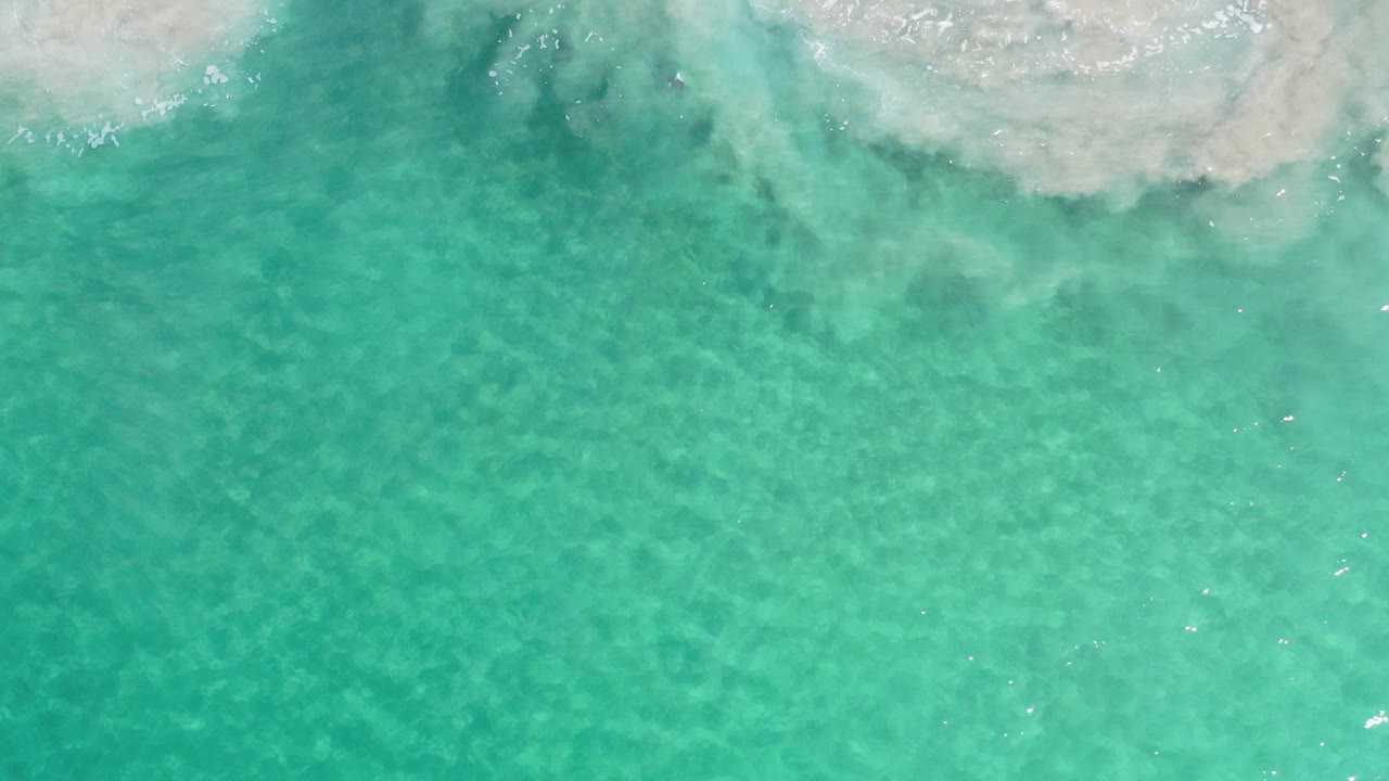 Overhead view of turquoise ocean in Los Cabos, Mexico, with white surf foam entering along the upper edge and no visible people.