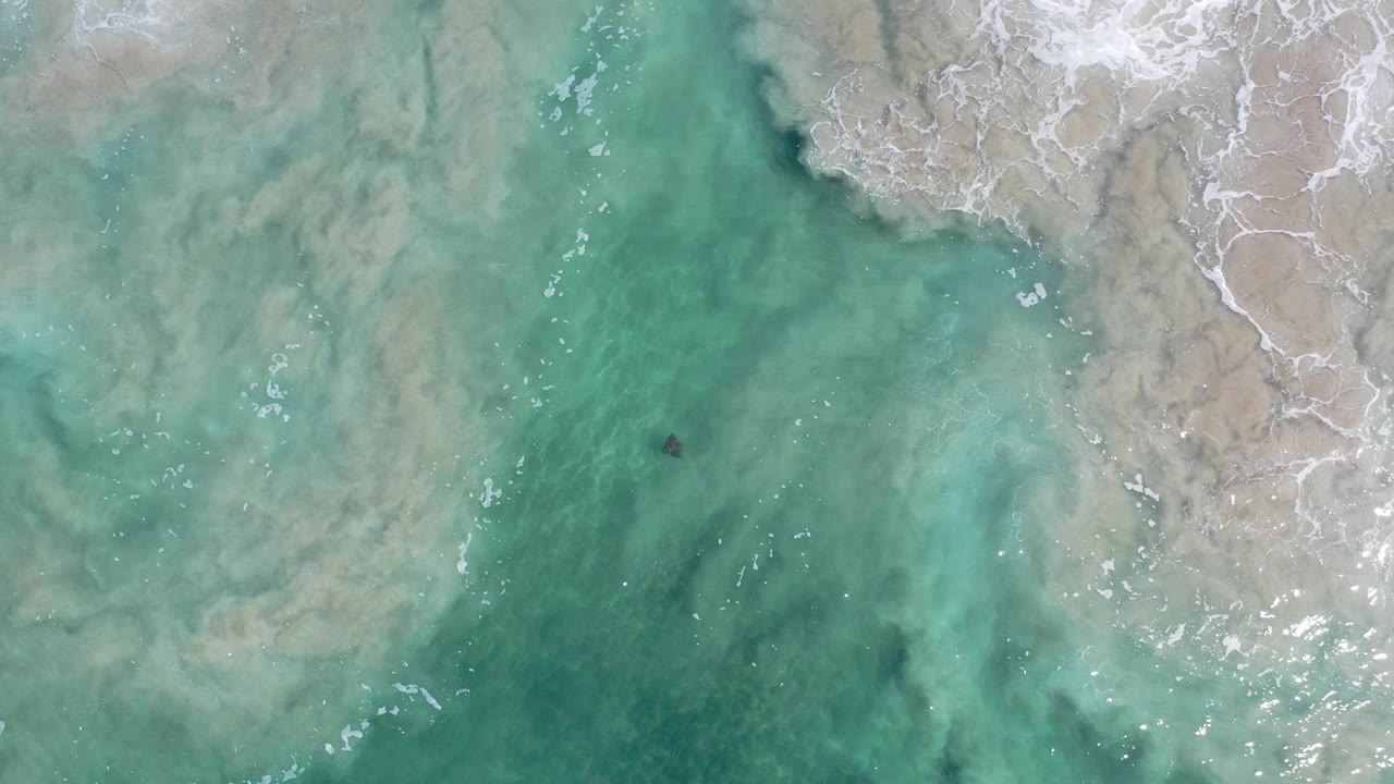 Aerial view of clear shallow water in Los Cabos, Mexico, with a pale sand shelf on the right, thin white foam lines, and no visible people.