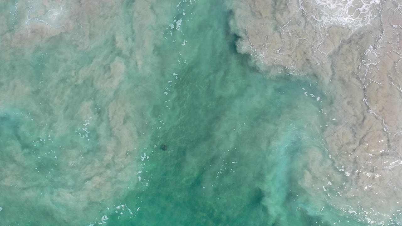 Overhead view of blue-green shallow water in Los Cabos with pale sand-toned swirls near the top edge, small surface ripples, and no visible people.