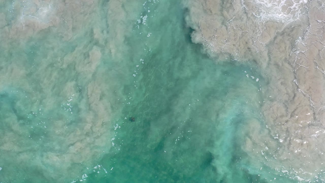 Overhead Los Cabos water frame with cloudy pale seabed patterns crossing the upper right, green-blue shallows below, scattered white ripples, and no visible people.