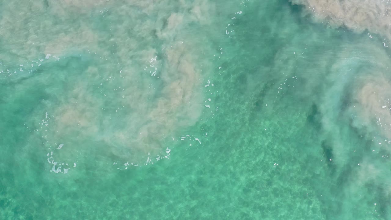 Blue-green ocean seen from above in Los Cabos, Mexico, with a cloudy white wave plume spreading across the upper right and no visible people.