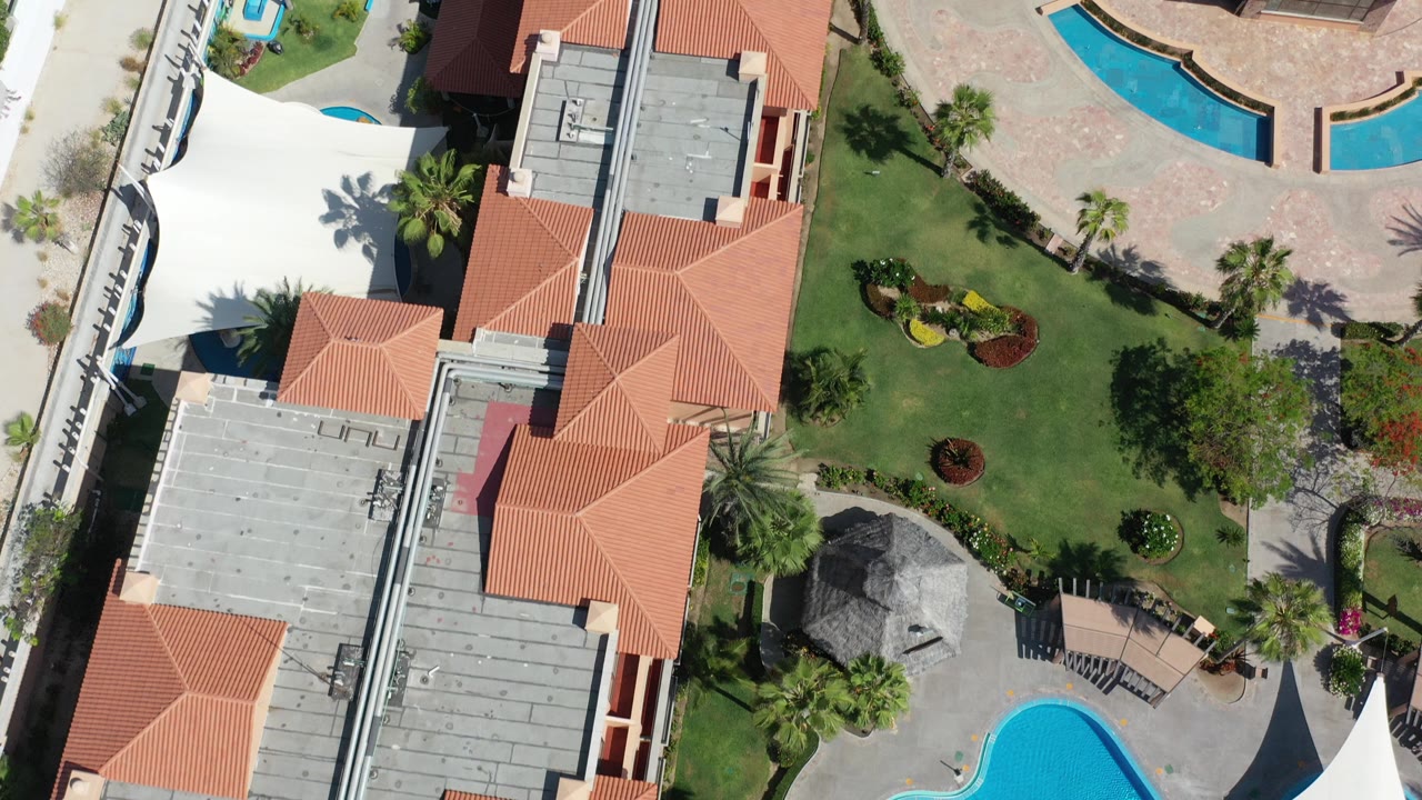 Overhead Los Cabos view of terracotta-roof buildings, landscaped lawns, palm trees, curved blue swimming pools, and pale paving with no visible people.