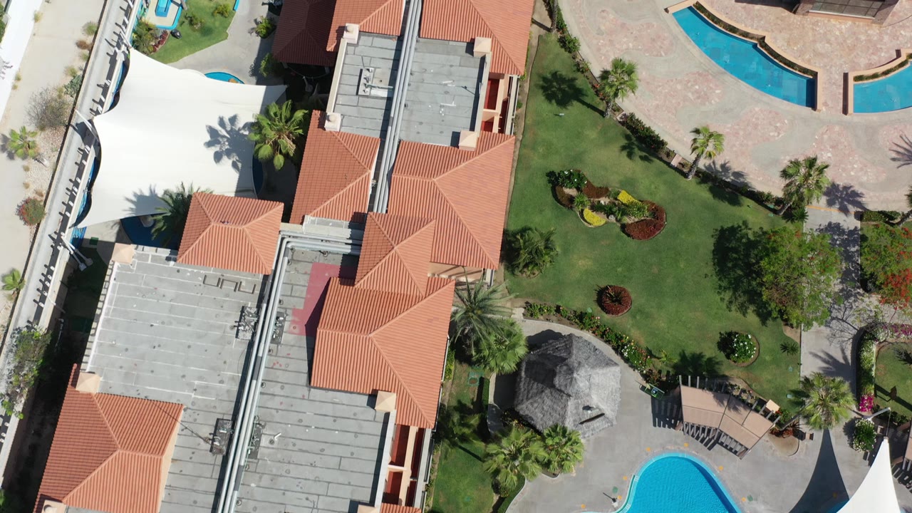Top-down Los Cabos frame with red tile roofs, gray rooftop surfaces, green landscaping, palm shadows, and bright blue pool shapes with no visible people.