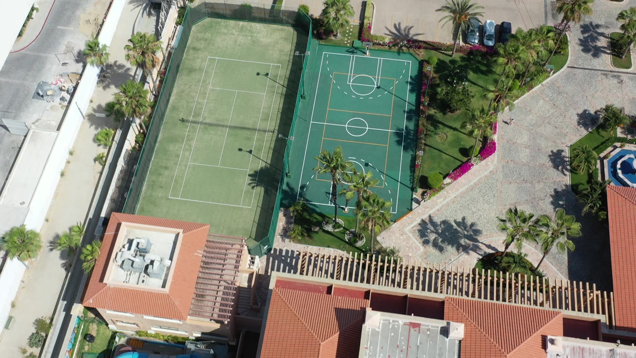 Overhead view of tennis and basketball courts beside parking and red tile roofs.