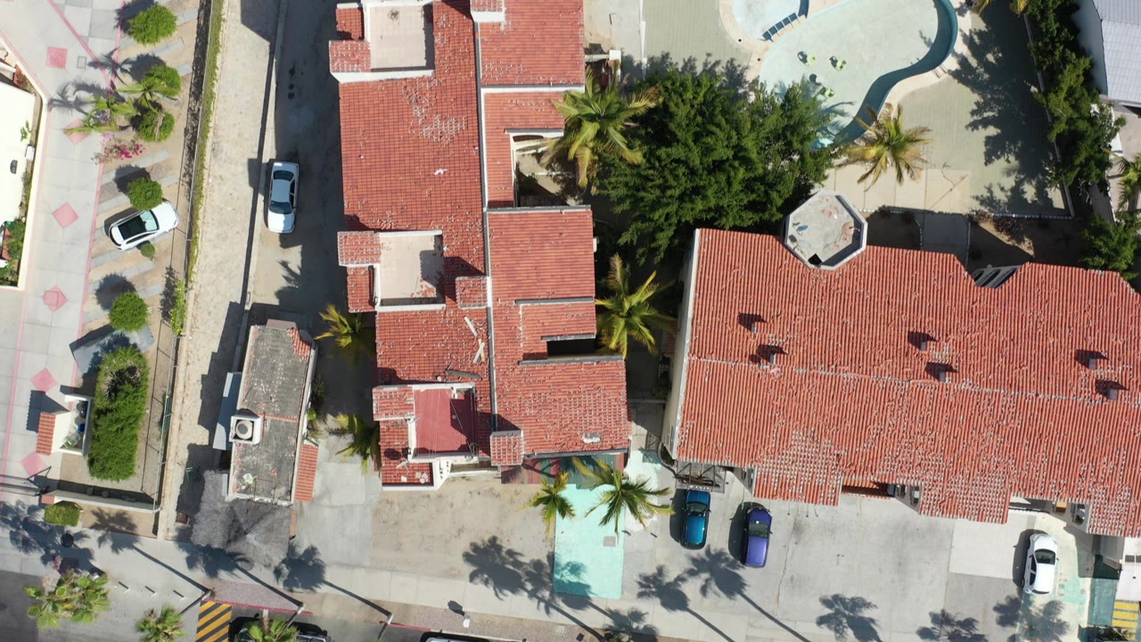 Top-down aerial frame in Los Cabos, Mexico showing red tile roof villas, a narrow residential street, parked cars, palm trees, and a small turquoise pool with no visible people.