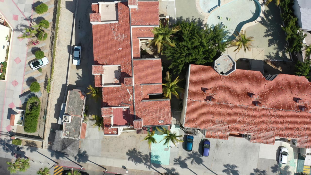 High overhead Los Cabos frame with red roof homes, palm trees, driveway parking, a pale curved pool area at the top right, and no visible people.