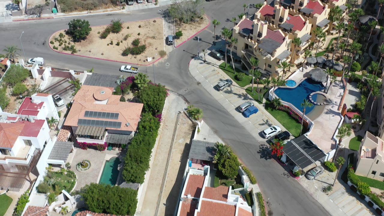 Slightly shifted top-down Los Cabos view showing a curved road between homes, turquoise pools, palms, parked vehicles, dry ground, and no visible people.