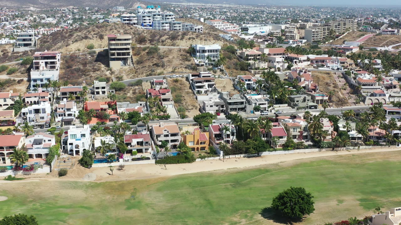 Opening aerial frame over Los Cabos, Mexico, with a green field in the foreground, palm-lined homes and a road in the middle, hillside buildings behind, and no visible people.