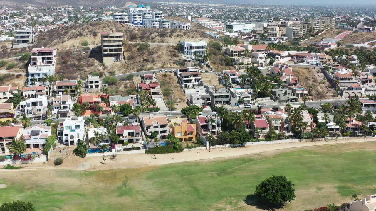 Los Cabos aerial view showing a road across the frame, clustered houses with red and white roofs, dry hillside lots, distant city blocks, and no visible people.