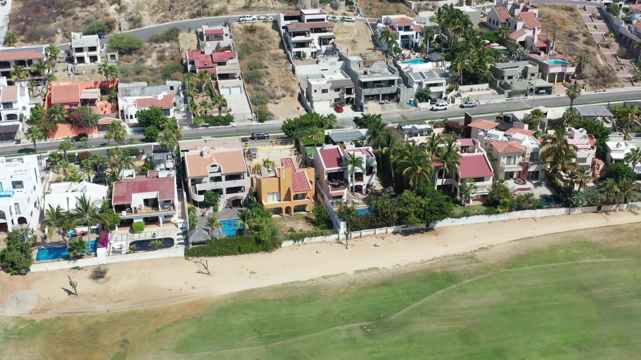 Aerial view of coastal homes, pools, road, sand, and green water.
