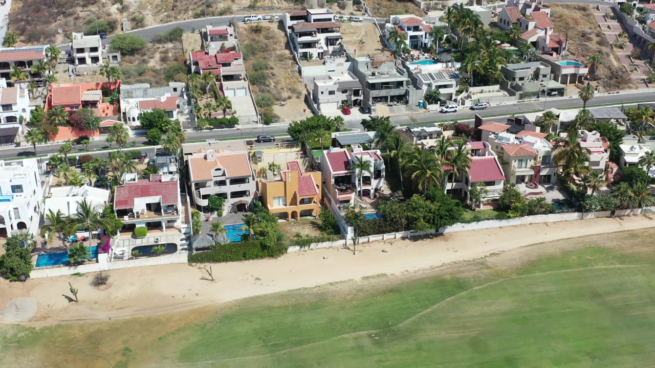 Aerial frame in Los Cabos, Mexico with red-roofed and white homes along the beach, blue pools inside walled properties, palms, a road, parked vehicles, and no visible people.